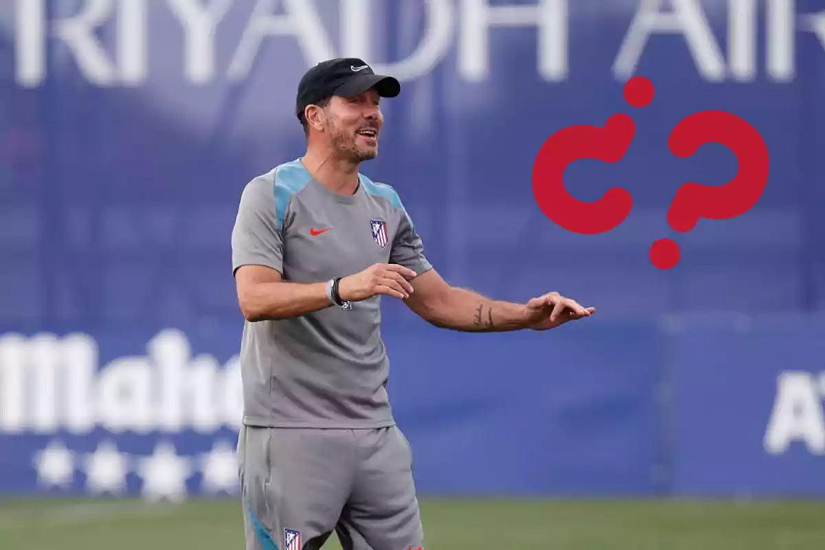 A football coach in a grey uniform and black cap gestures during a training session on a football field.