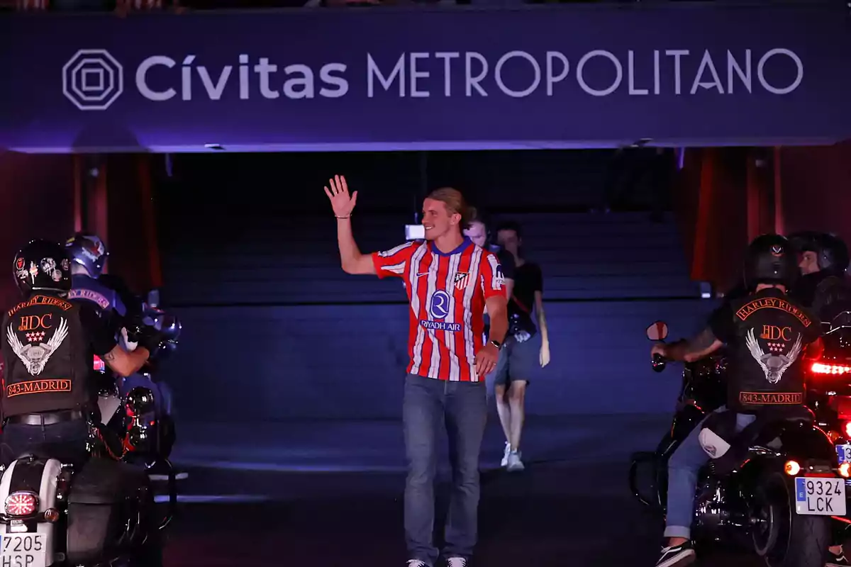 A man in a red and white striped shirt waves as he walks through a tunnel at the Cívitas Metropolitano stadium, surrounded by motorcyclists.