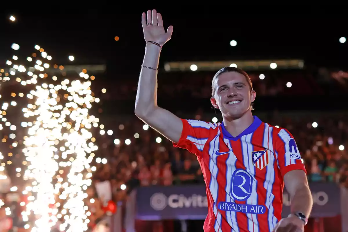 A football player wearing an Atlético de Madrid shirt greets fans in a stadium illuminated with fireworks and backlighting.