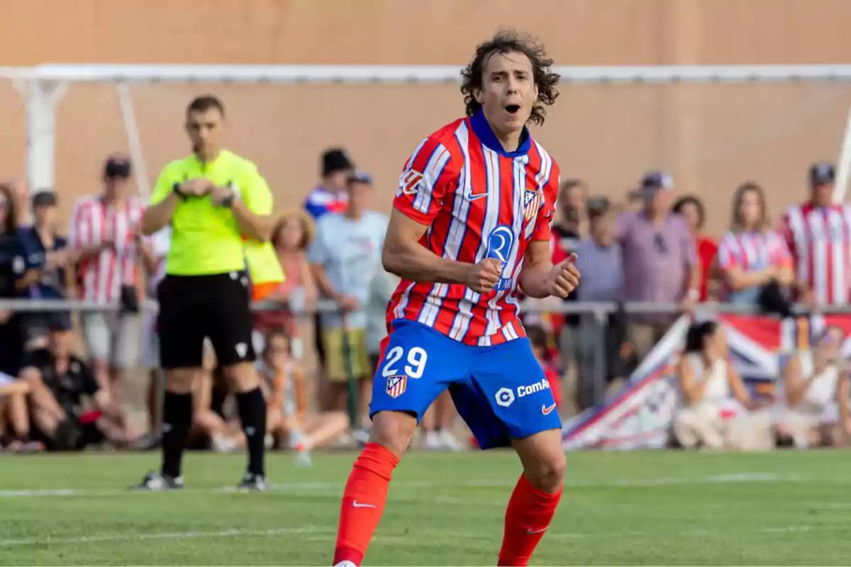 Soccer player in red and white uniform celebrating on the field with a referee and spectators in the background.