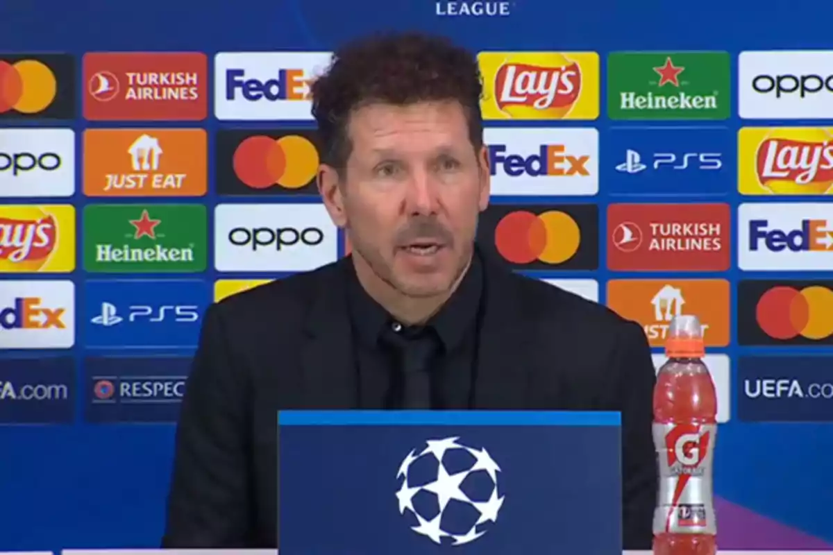 A man speaking at a UEFA Champions League press conference with various sponsor logos in the background.