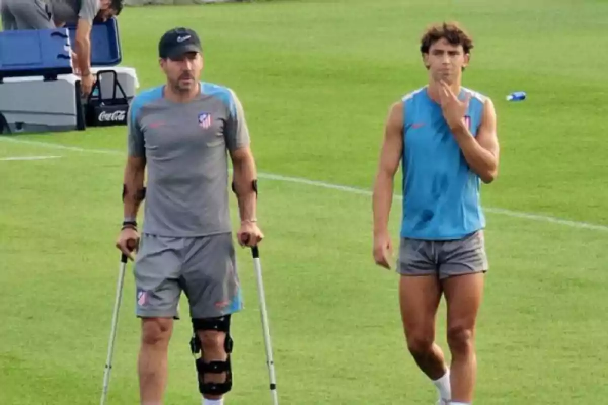 Two people on a soccer field, one of them with crutches and a knee brace, both wearing Atlético de Madrid sports clothing.