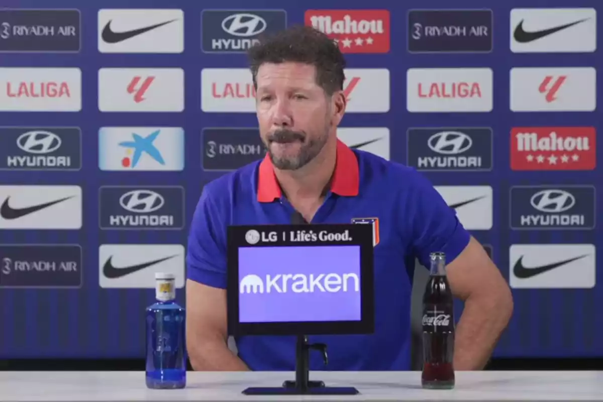 A man with a beard and moustache, wearing a blue T-shirt with a red collar, is sitting at a table during a press conference. In front of him are a bottle of water and a bottle of Coca-Cola. In the background are various logos of sponsors such as Hyundai, Nike, Mahou and LaLiga.