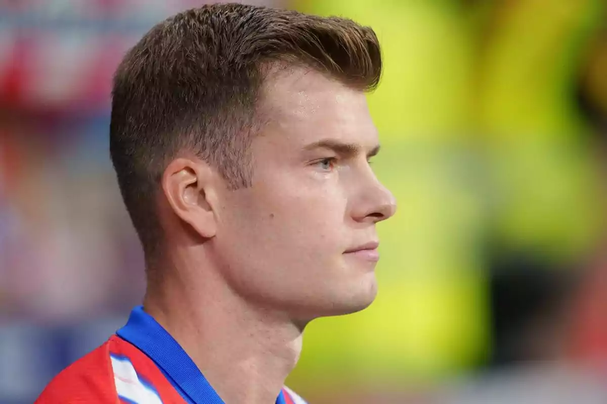 A man in a red and blue t-shirt looks to the right with a serious expression and an out-of-focus background.