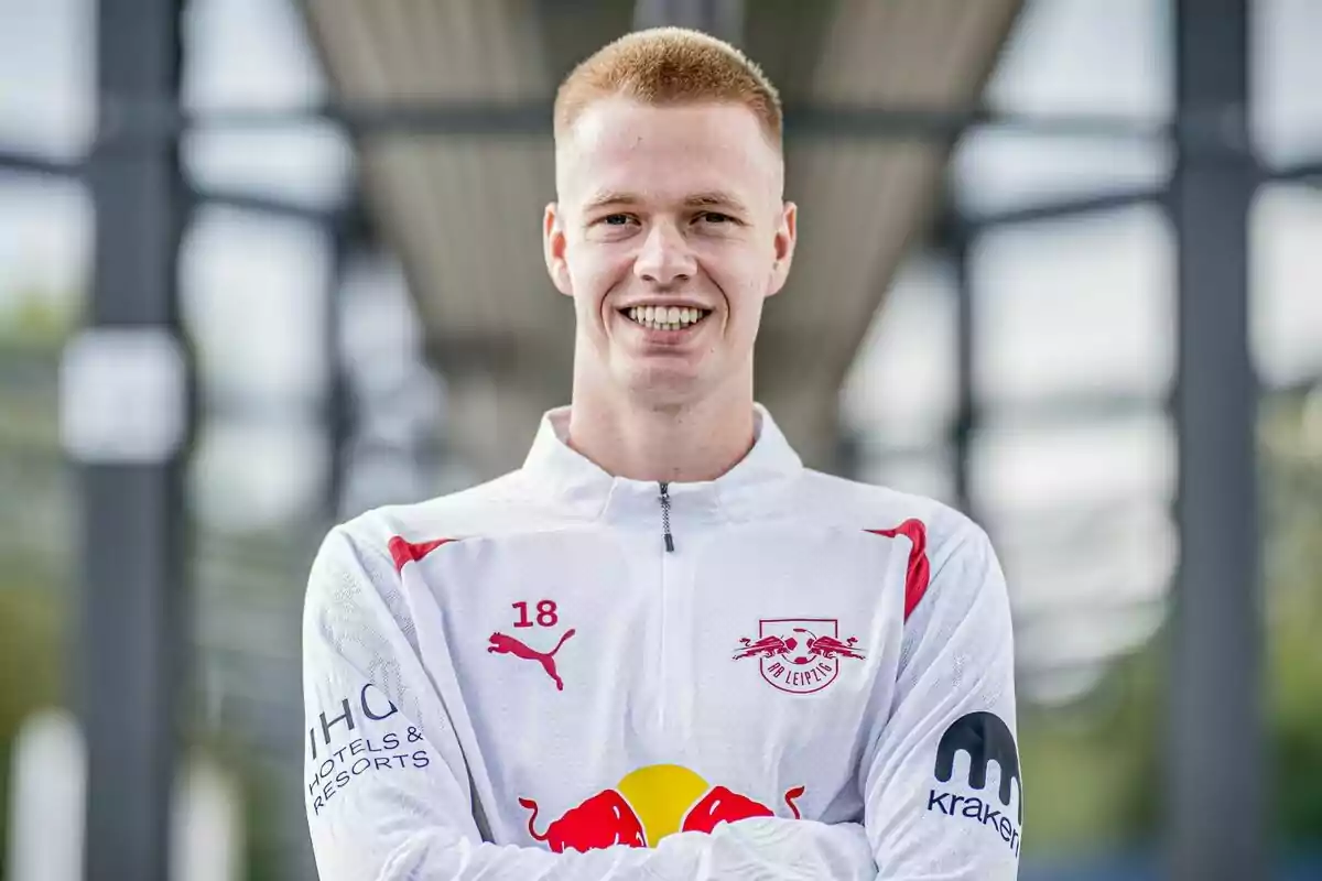 A young man in the uniform of the RB Leipzig football team smiles with his arms crossed.