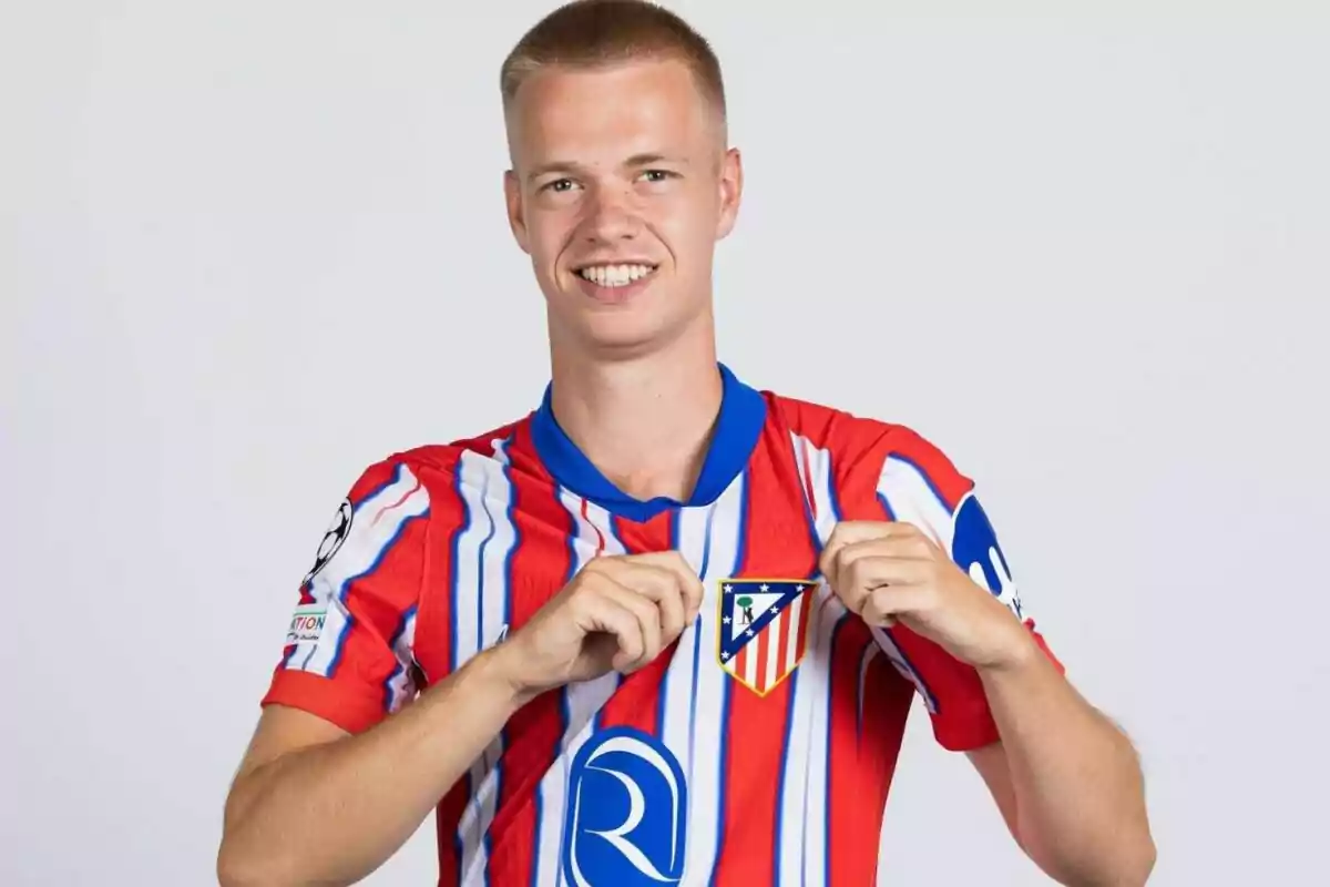 A soccer player in a red and white jersey holds up his team's crest while smiling at the camera.