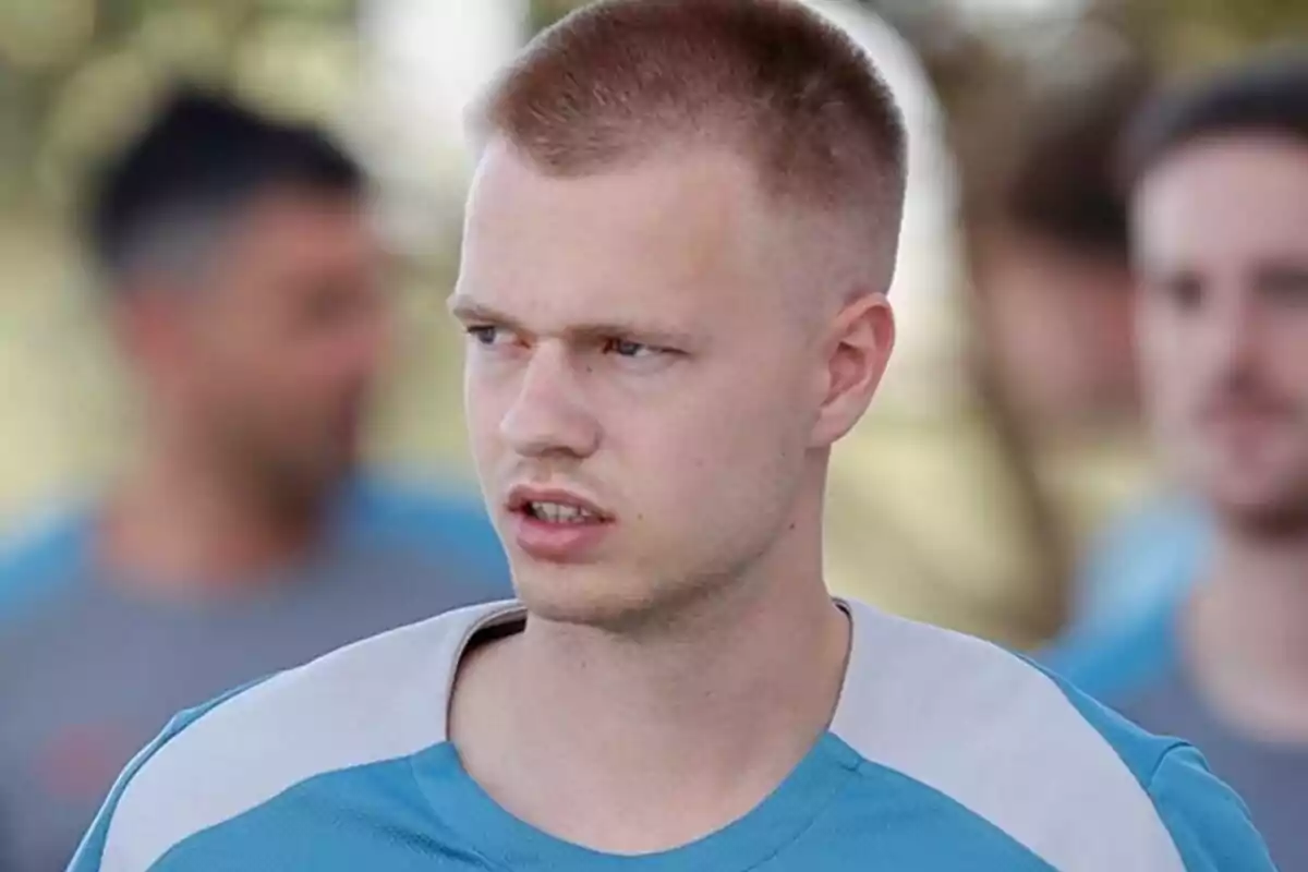 A man in a light blue t-shirt and short haircut, looking to the right with a serious expression, with people out of focus in the background.