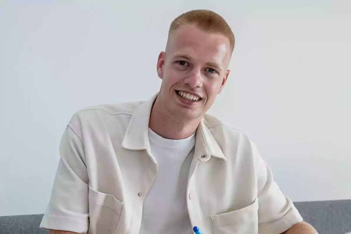 A young man with short blond hair, smiling and wearing a beige shirt over a white t-shirt, sitting on a grey sofa with a white wall in the background.