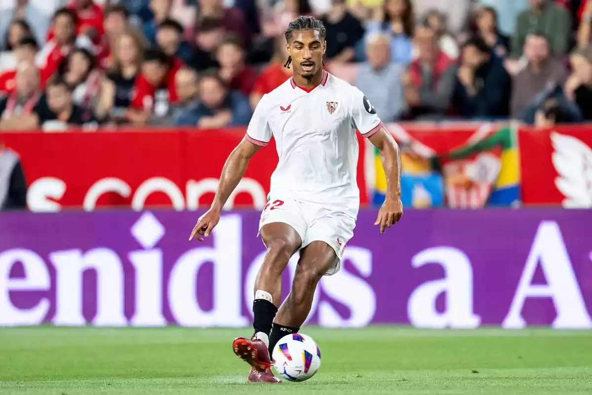 A Sevilla FC soccer player, wearing the team's white uniform, controls the ball on the field during a match, with the crowd of spectators in the stands in the background.