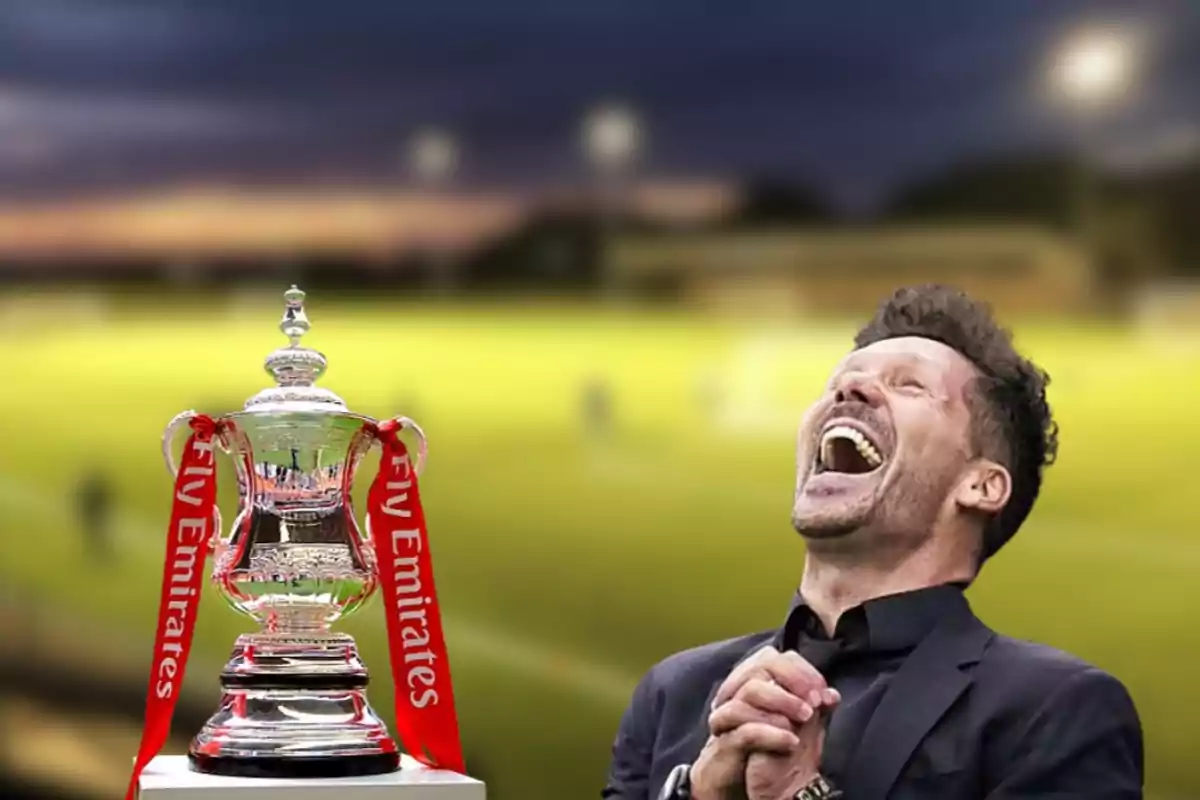 Man laughing next to a football trophy with red ribbons saying "Fly Emirates" on a blurred football field at sunset.