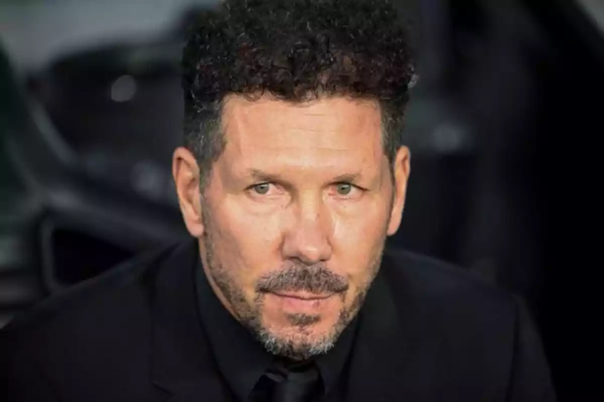 Man with curly hair and beard, dressed in black suit, looking forward.