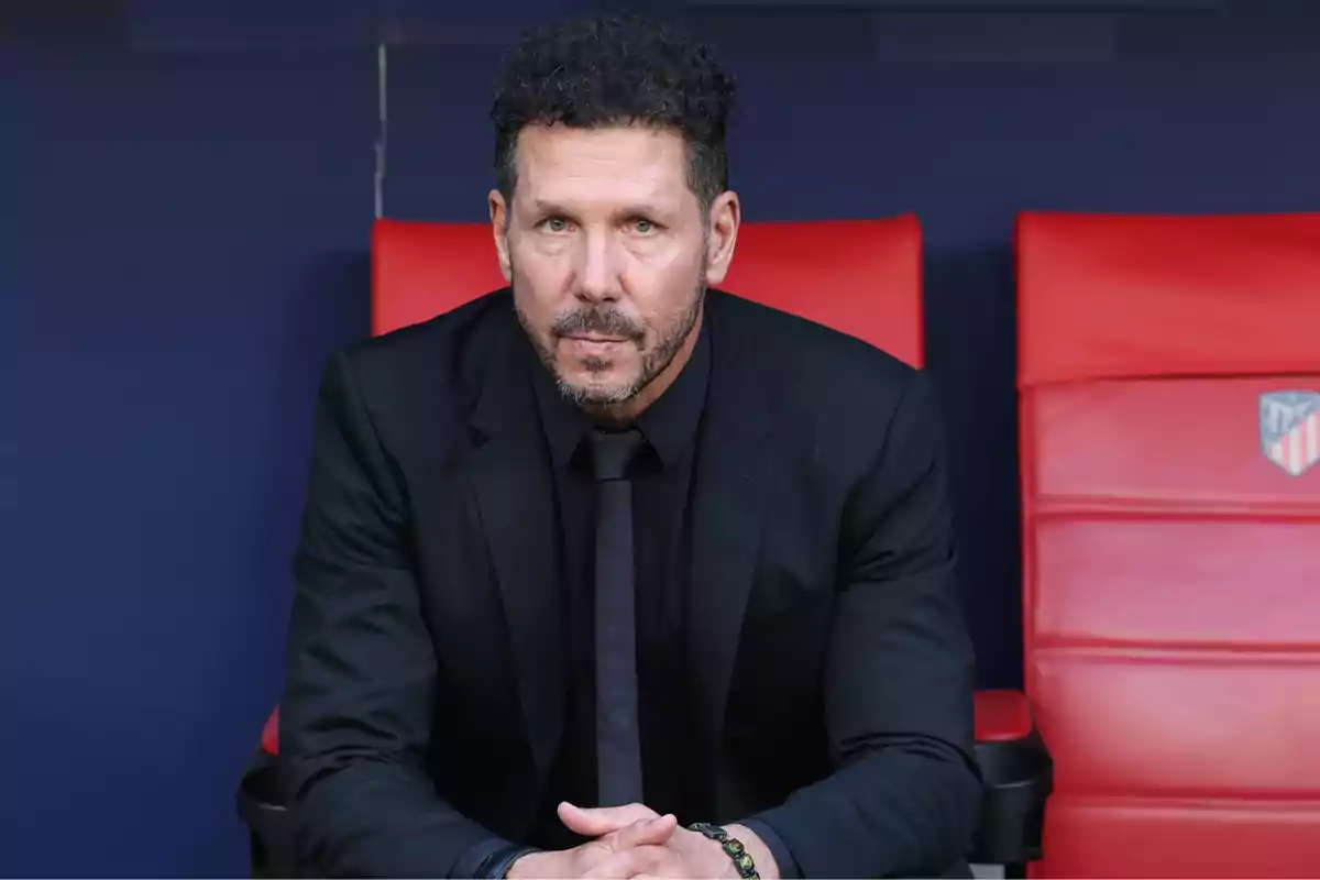 Man in a black suit sitting on a sports bench with red seats.