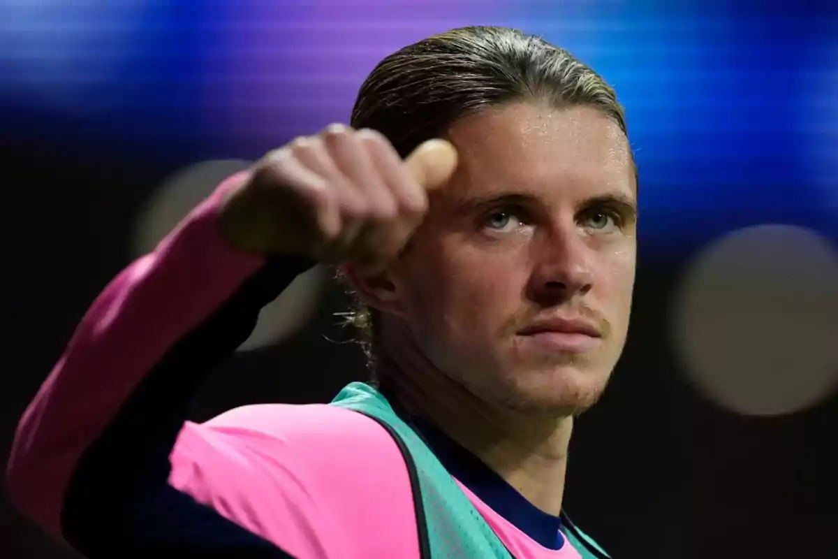 A soccer player in a pink and green uniform raises his fist in a sign of determination against a blurred background of blue lights.