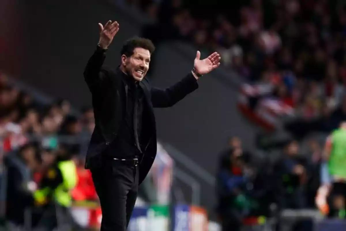 Person dressed in black gesturing with raised arms in a stadium.