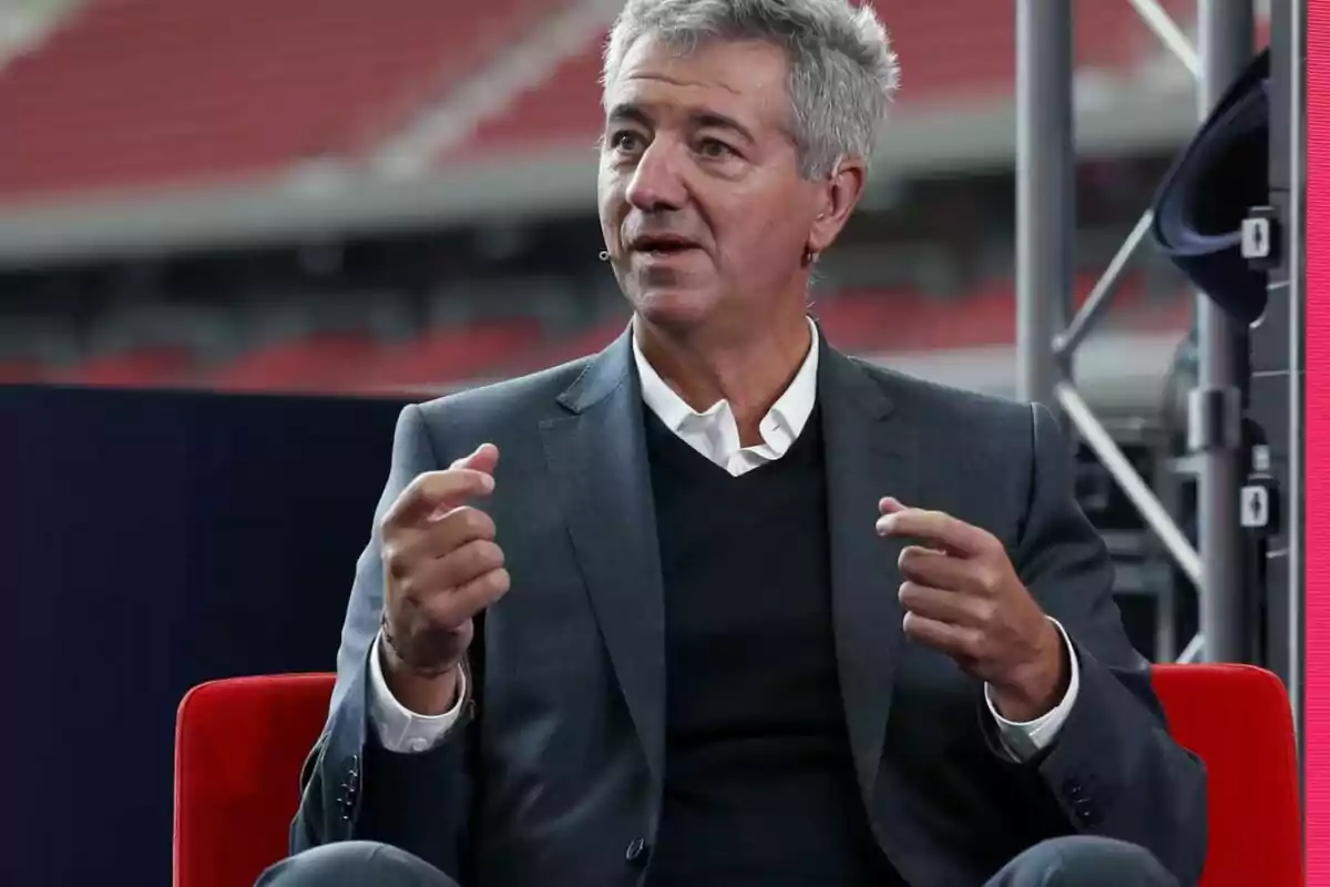 Gray-haired man in a dark suit and white shirt speaking on a stage with an out-of-focus background.