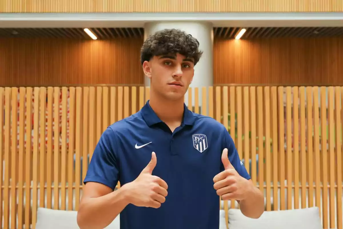 A young man in a blue Atletico Madrid shirt gives the thumbs up gesture in front of a wooden slatted wall.