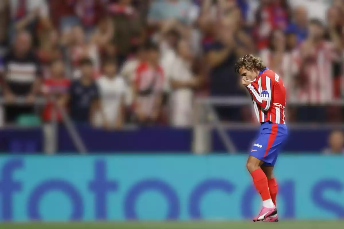 A soccer player in a red and white uniform kisses his jersey on the field as the crowd in the stands watches.