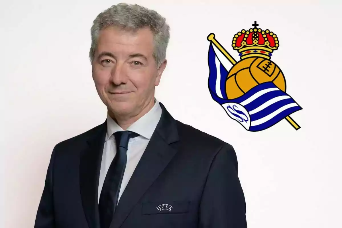 A man in a suit and tie, with the UEFA logo in his pocket, posing in front of a white background with the Real Sociedad crest.