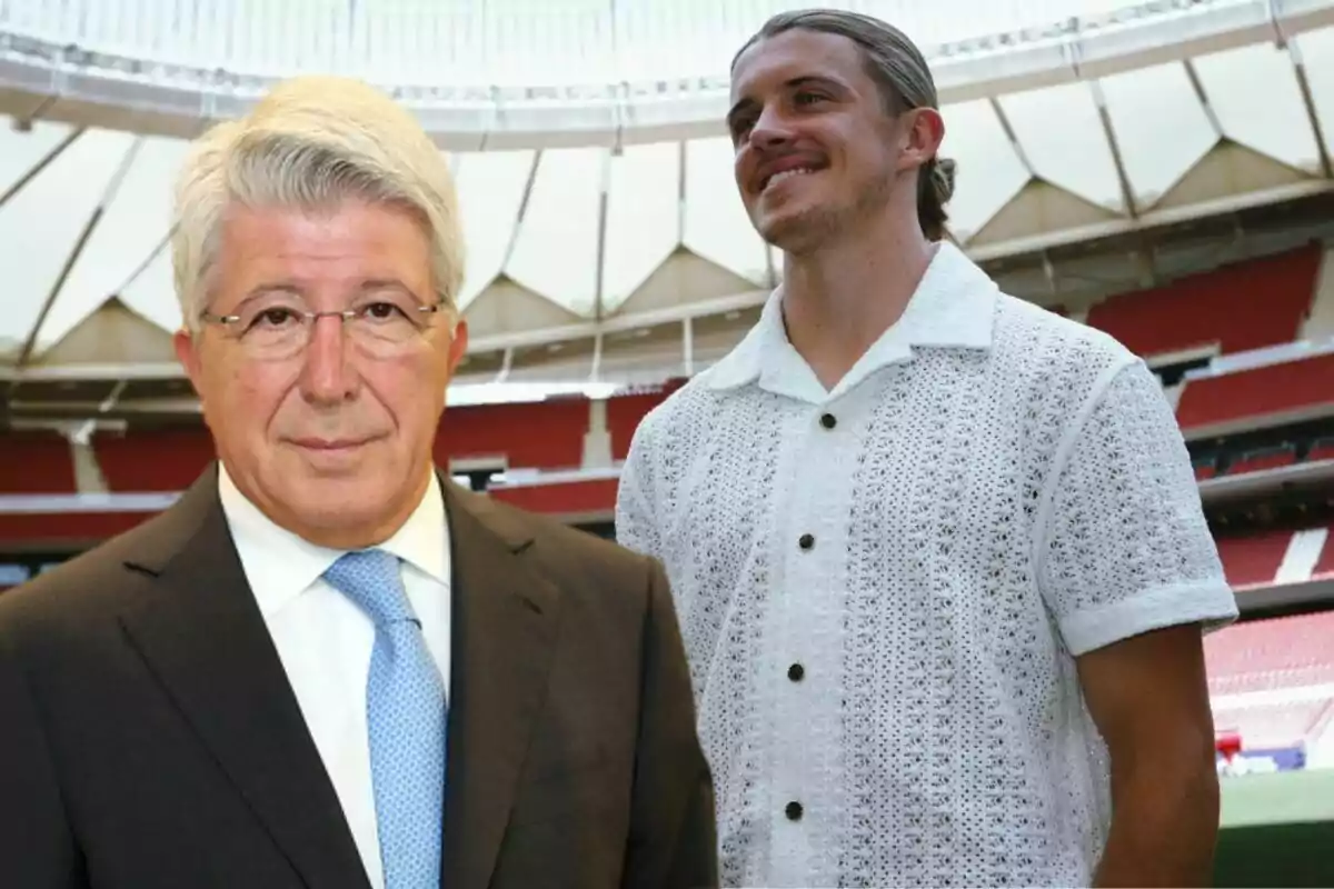 Two men pose in a stadium, one of them wearing a suit and tie, while the other wears a white knitted shirt.