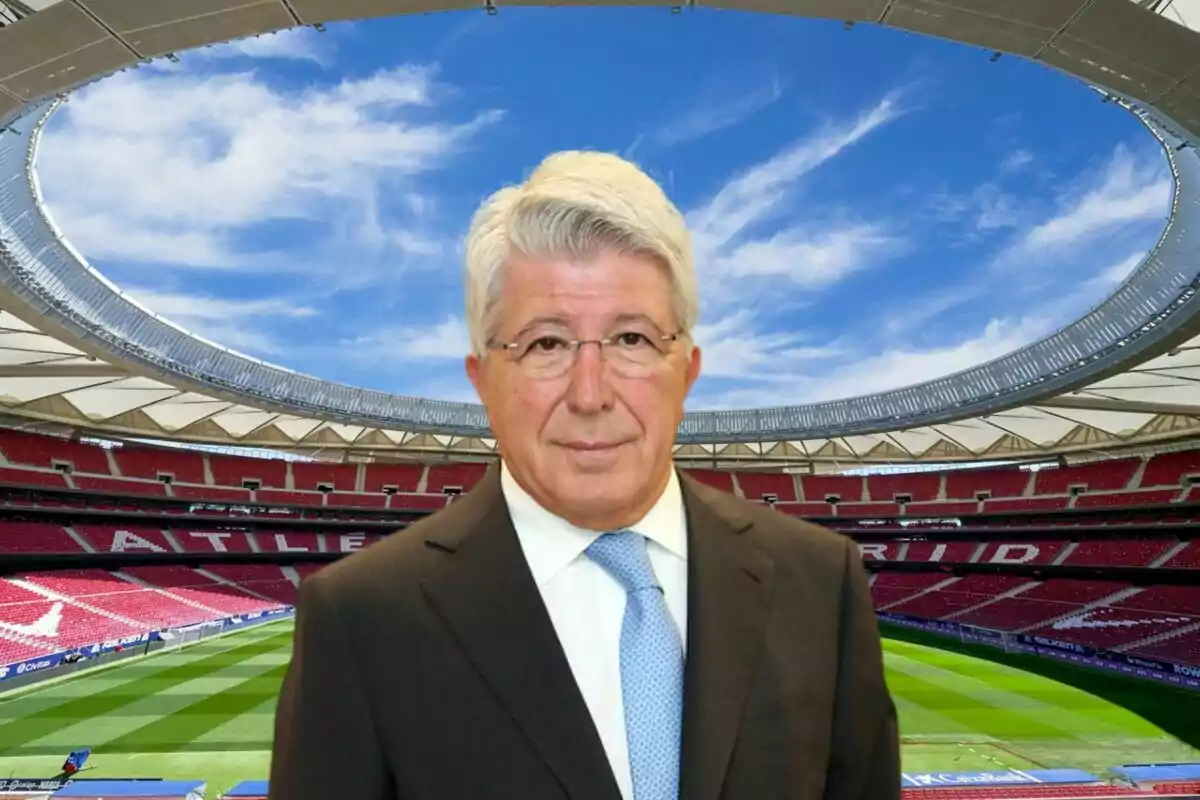 A man with grey hair and glasses, dressed in a suit and tie, posing in a football stadium with red stands and a blue sky in the background.