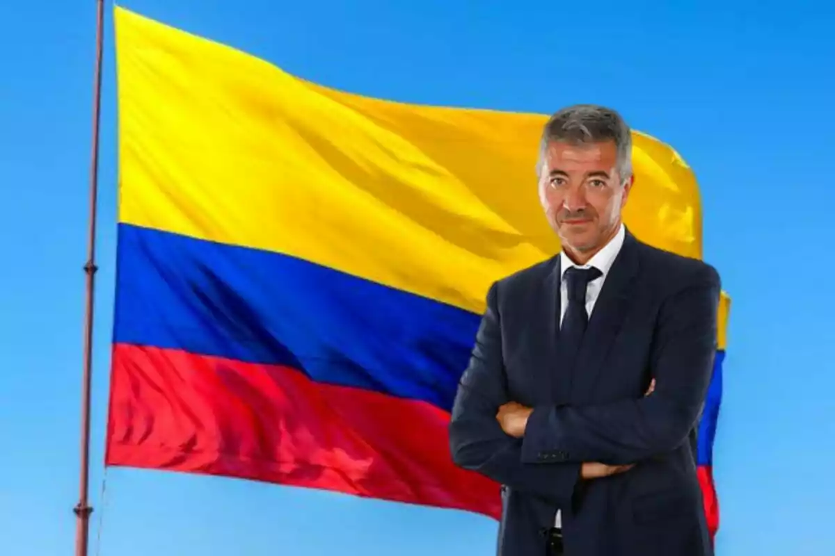 A man in a dark suit with his arms crossed in front of a waving Colombian flag.