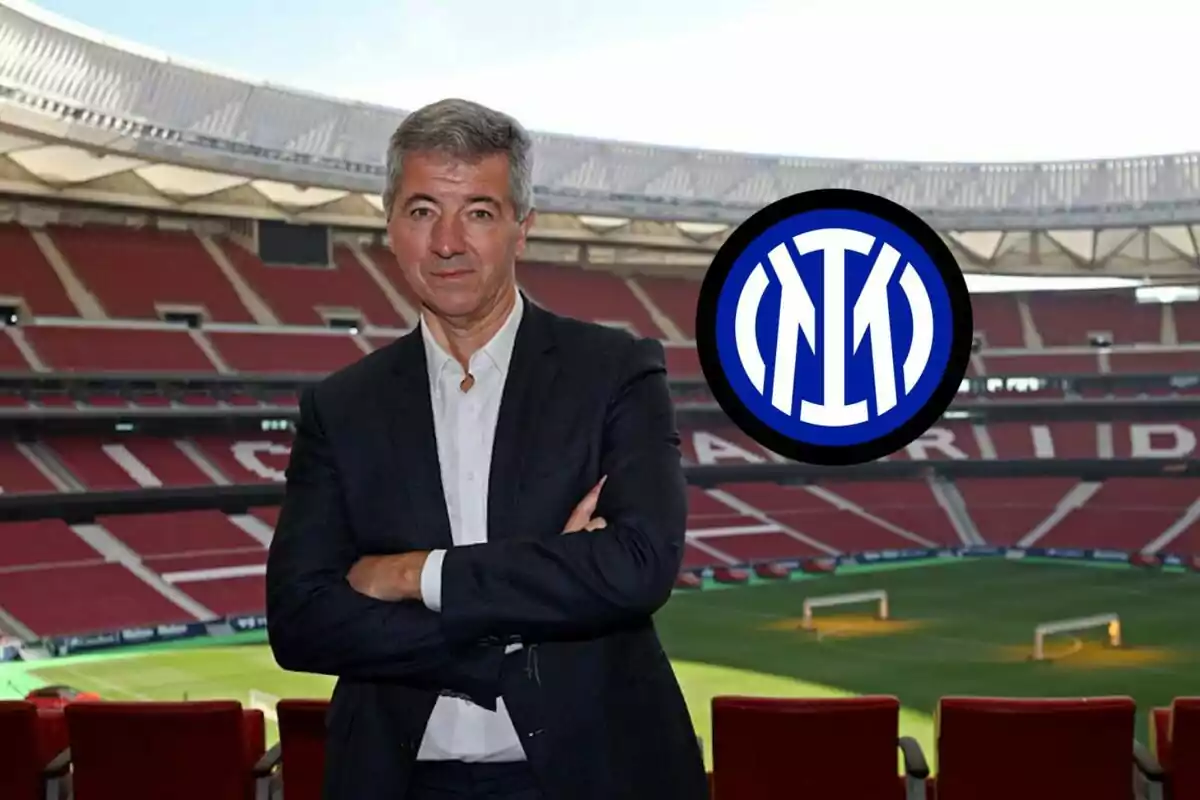 A man in a dark suit with his arms crossed stands in an empty football stadium with red seats and a modern roof, next to an Inter Milan logo.