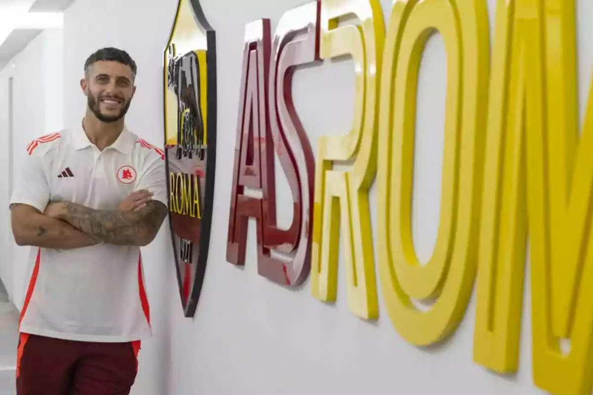 A man in a white and red T-shirt stands next to a large sign reading "AS ROMA" in a brightly lit hallway.