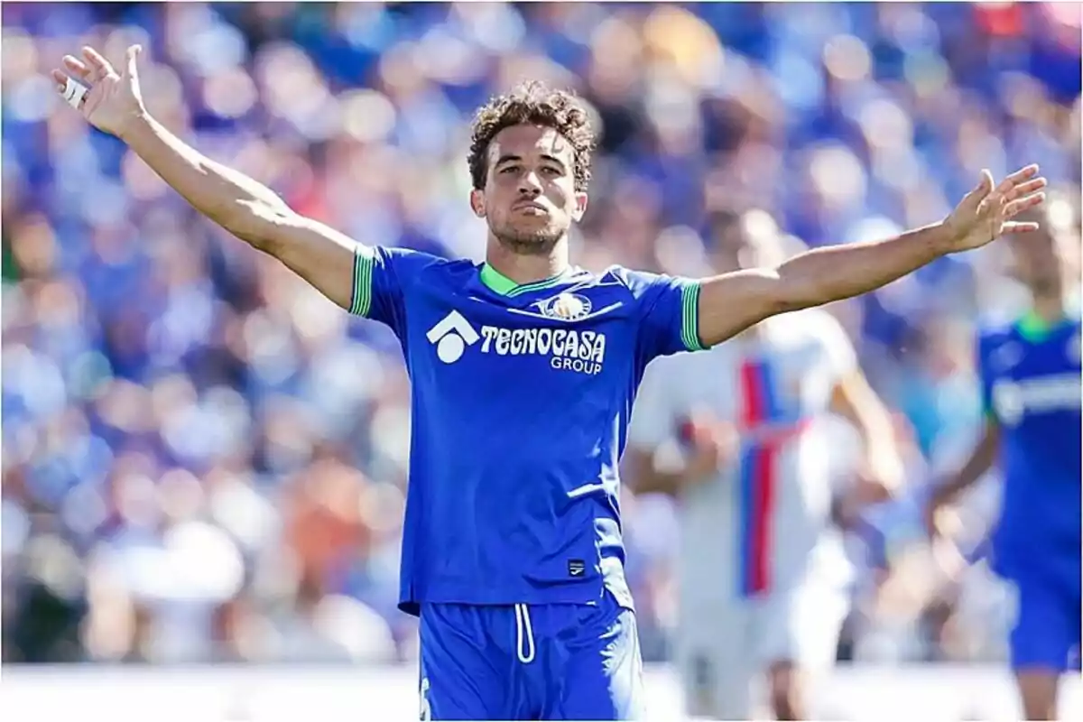 Soccer player in blue uniform celebrating with arms outstretched in a stadium.