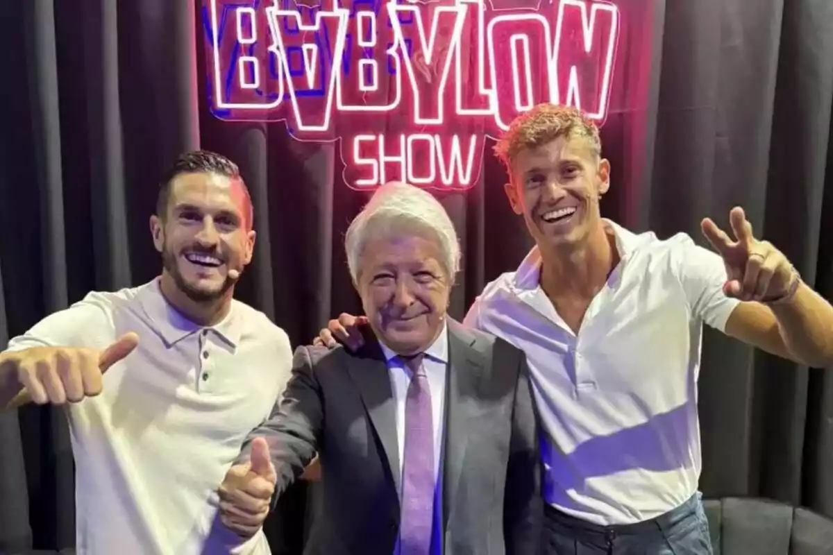 Three smiling men pose in front of a neon sign that reads 