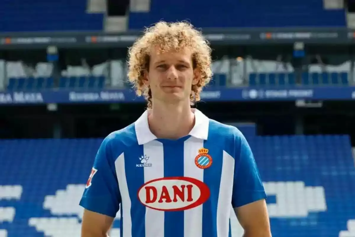 A soccer player with curly blond hair wears the blue and white uniform of RCD Espanyol in an empty stadium.