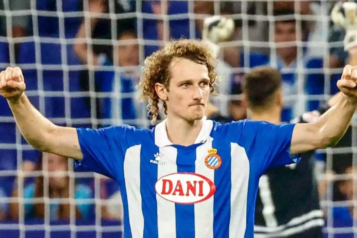 Soccer player in blue and white uniform celebrating with arms raised in front of a goal.