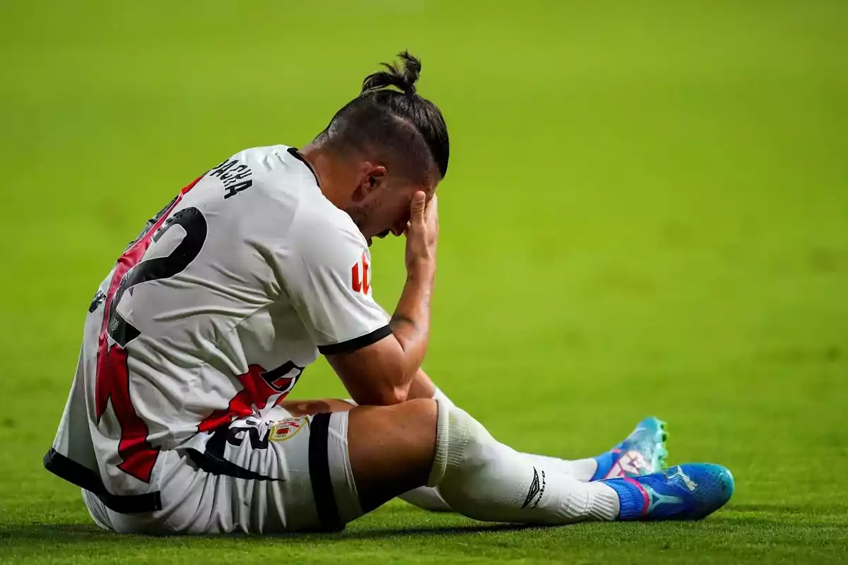 Soccer player sitting on field with hand on face showing frustration.