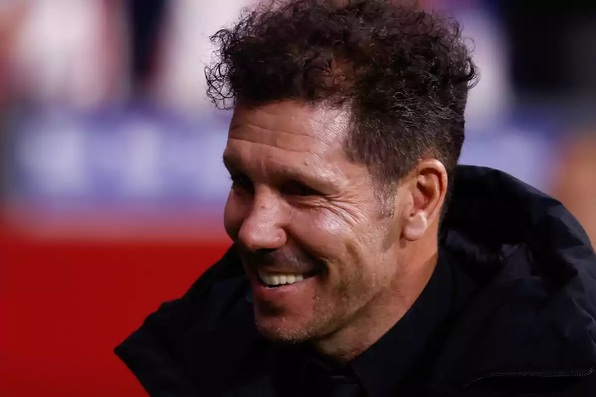 Smiling man with curly hair and light beard, wearing black jacket, in out of focus background.