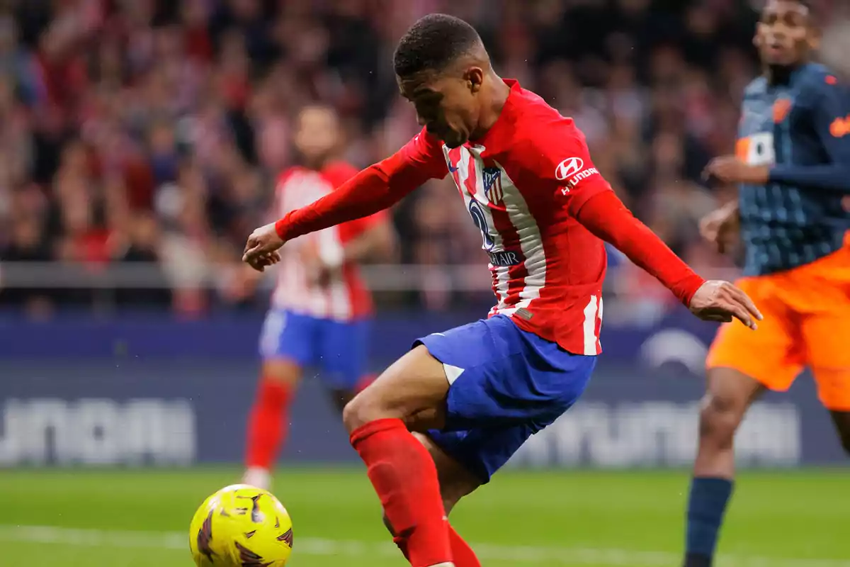 An Atletico Madrid soccer player is about to kick the ball during a match, while another player from the opposing team watches in the background.