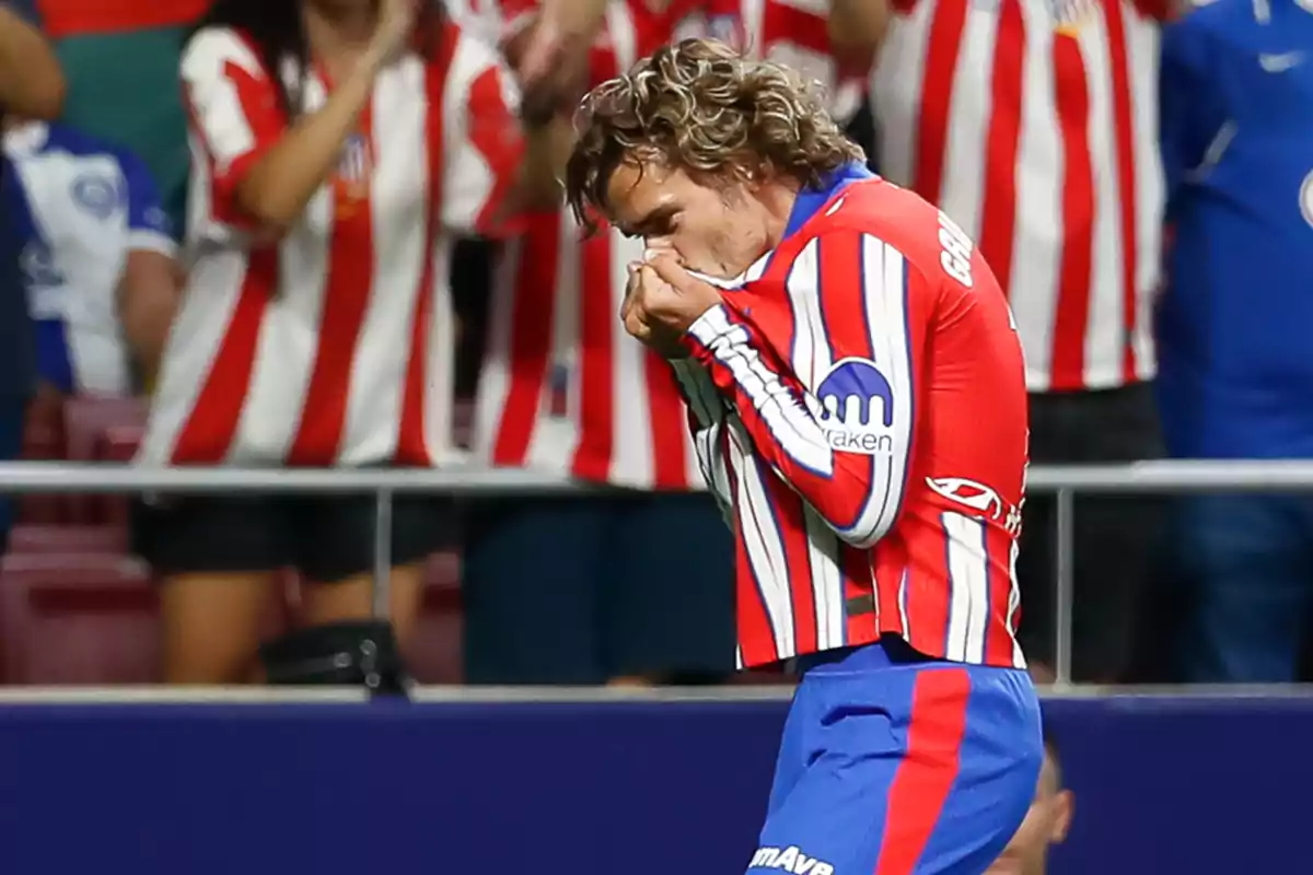 A soccer player kisses the crest on his shirt as he celebrates a goal, with fans in the background applauding and wearing red and white striped shirts.