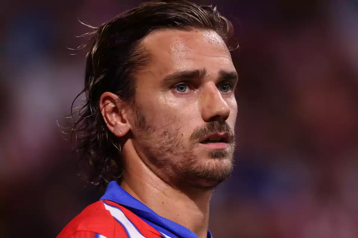 A soccer player in a red and blue jersey, with a serious expression and long hair, in a stadium.