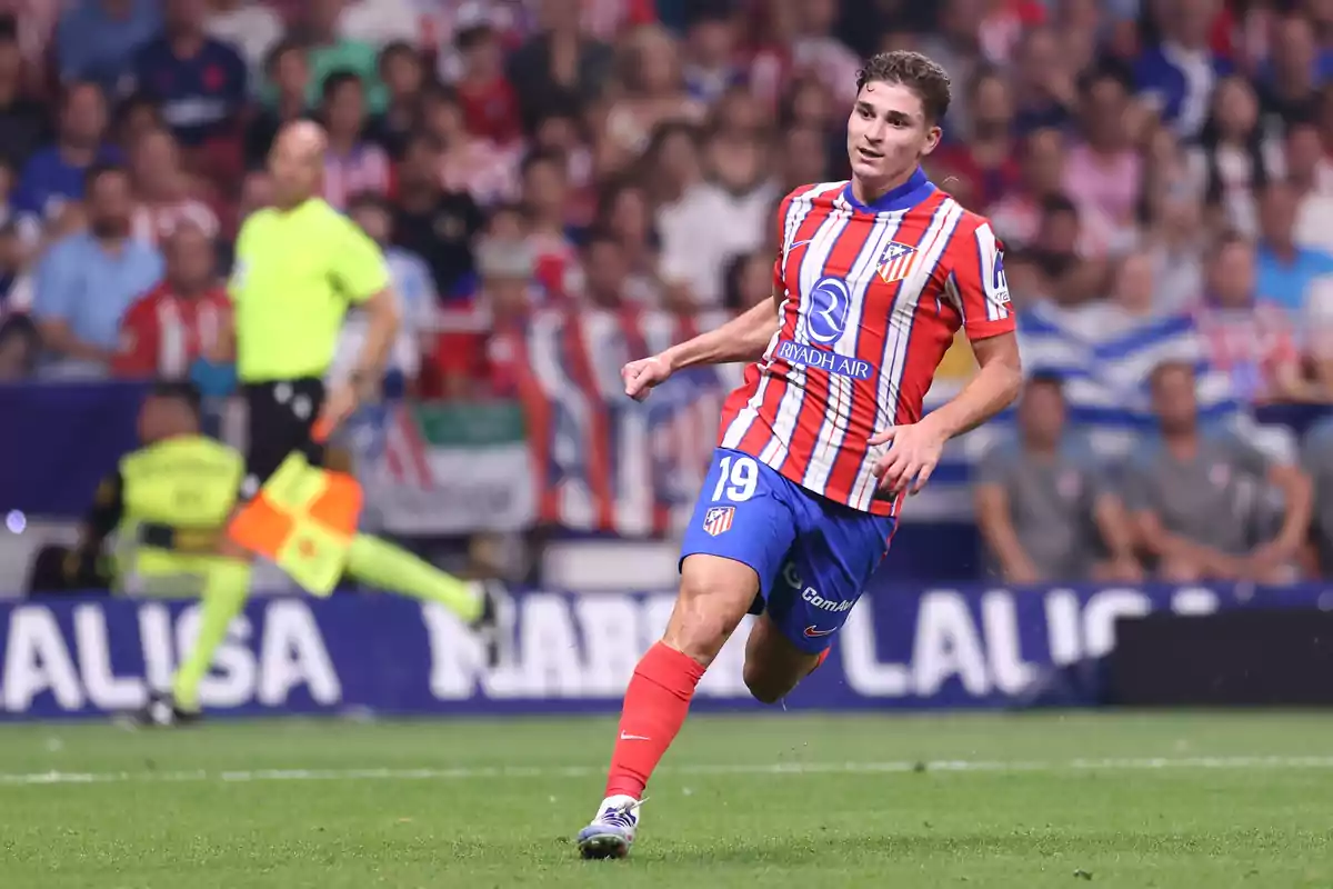 A football player in an Atletico Madrid uniform running on the pitch during a match, with a referee and spectators in the background.