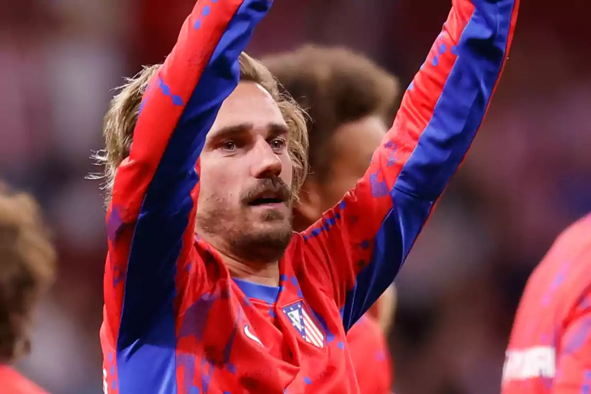 A soccer player in a red and blue uniform raises his arms in celebration.