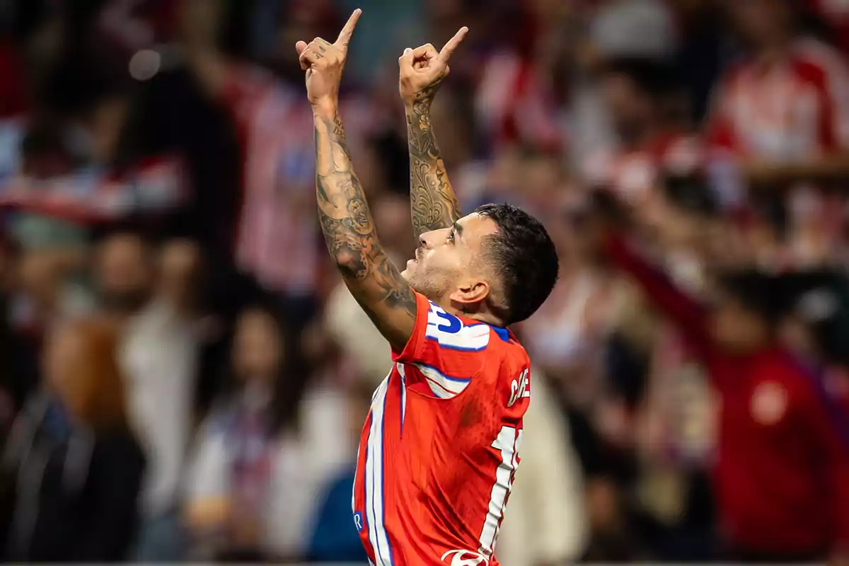 Soccer player celebrating with arms raised and pointing upwards, wearing a red, white and blue uniform, with tattoos visible on both arms and a stadium full of fans in the background.