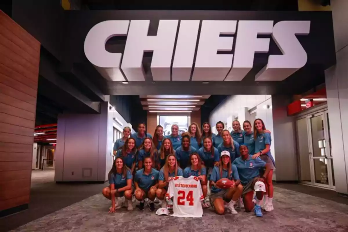 A group of women posing with a t-shirt and a football under a sign that says "CHIEFS."