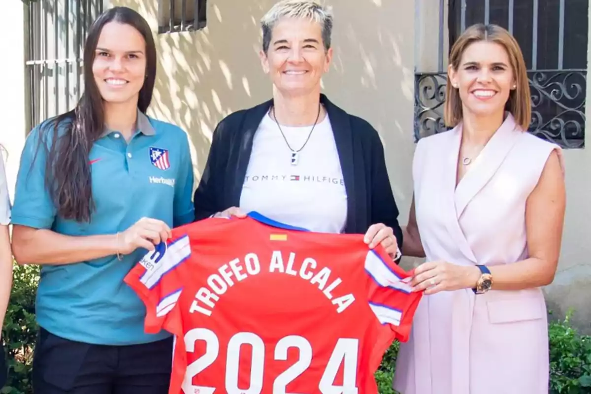 Three smiling women pose with a red t-shirt that says "ALCALA 2024 TROPHY."