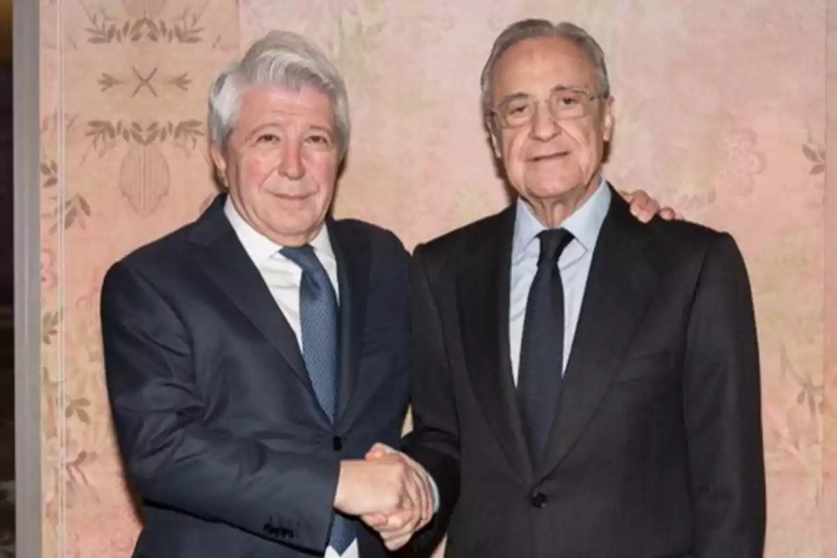 Two men in suits and ties shake hands and pose for the camera against a decorative wall in the background.