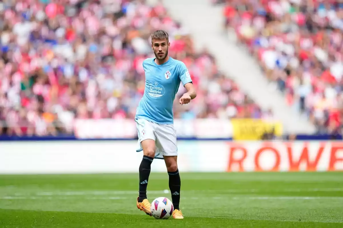 A soccer player in a light blue and white uniform controls the ball in a stadium full of spectators.