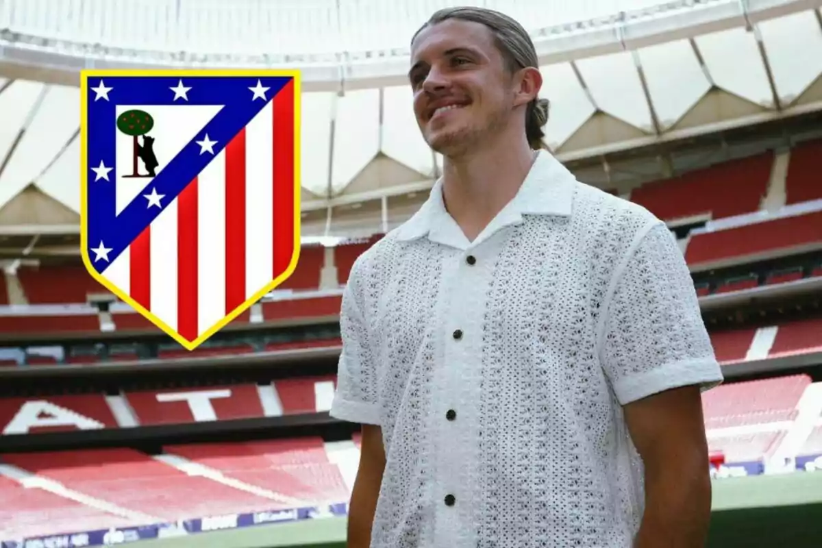 A smiling man in a white knitted shirt posing in a football stadium with the Atletico Madrid crest next to him.