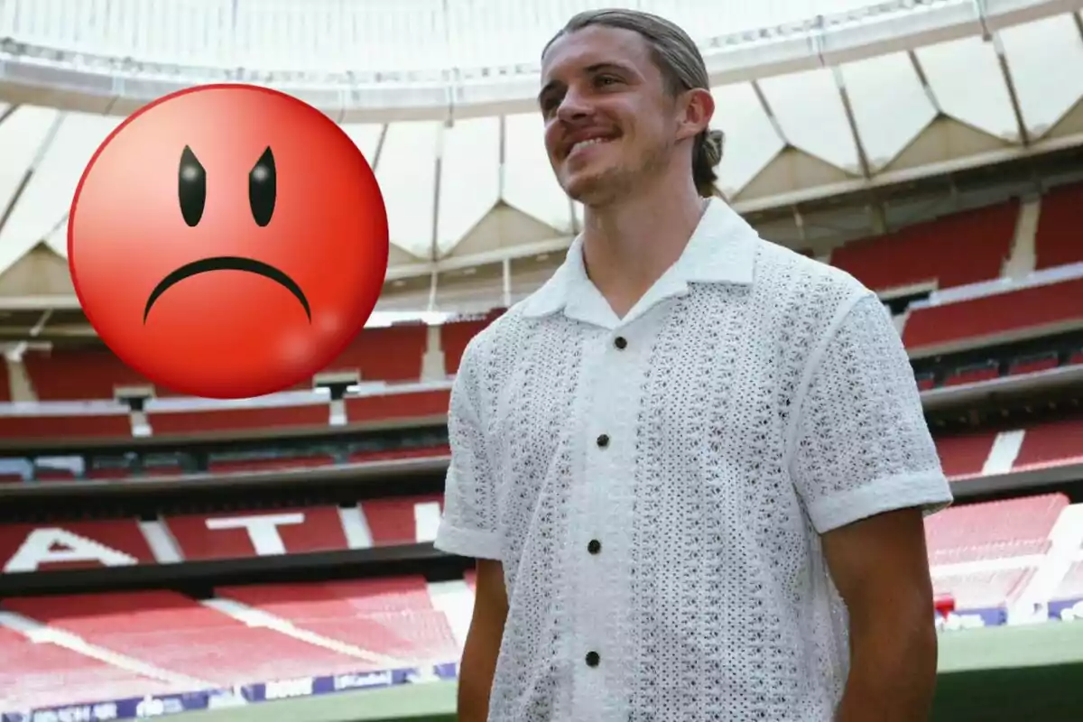 A smiling man in a white knitted shirt stands in a football stadium, while a large angry face emoji is superimposed on the image.