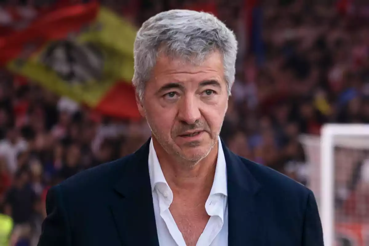 A man with gray hair and a stubble beard, dressed in a white shirt and a dark jacket, stands in front of a crowd with out-of-focus flags in the background.