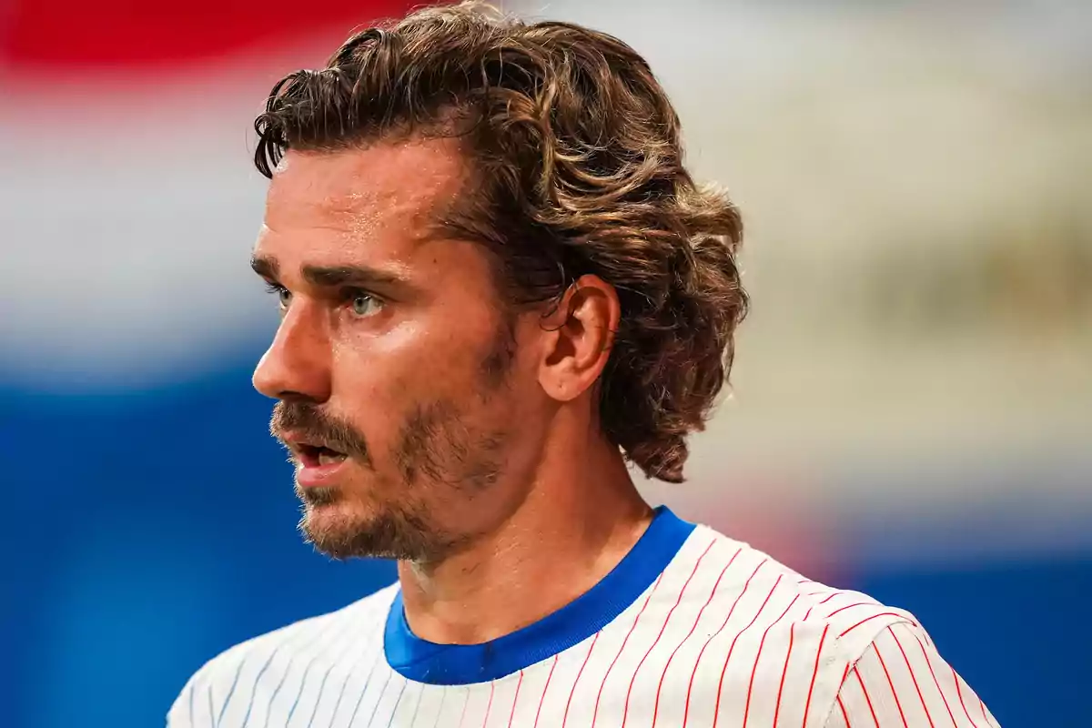 Man in a white sports shirt with red stripes and a blue collar, in profile, with curly hair and a beard, in a sports environment.