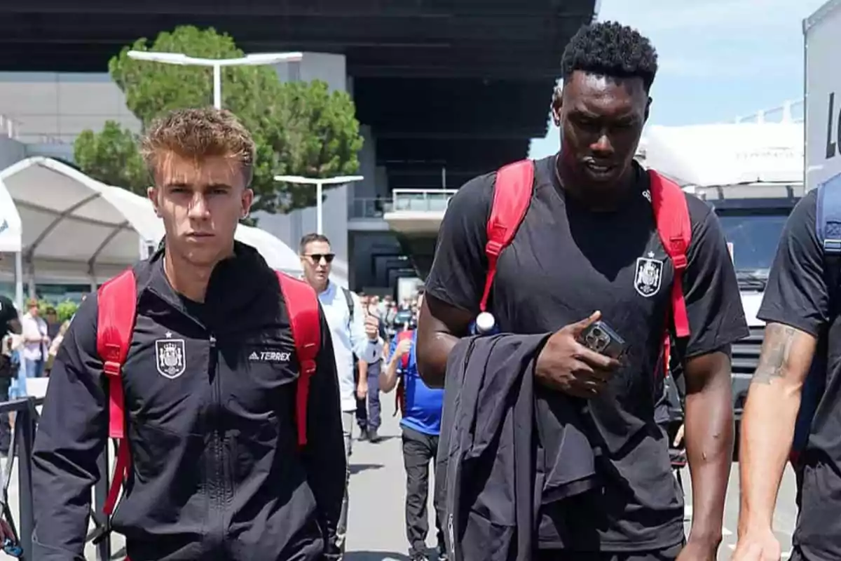 Two men with red backpacks walk outdoors, one of them holding a mobile phone.