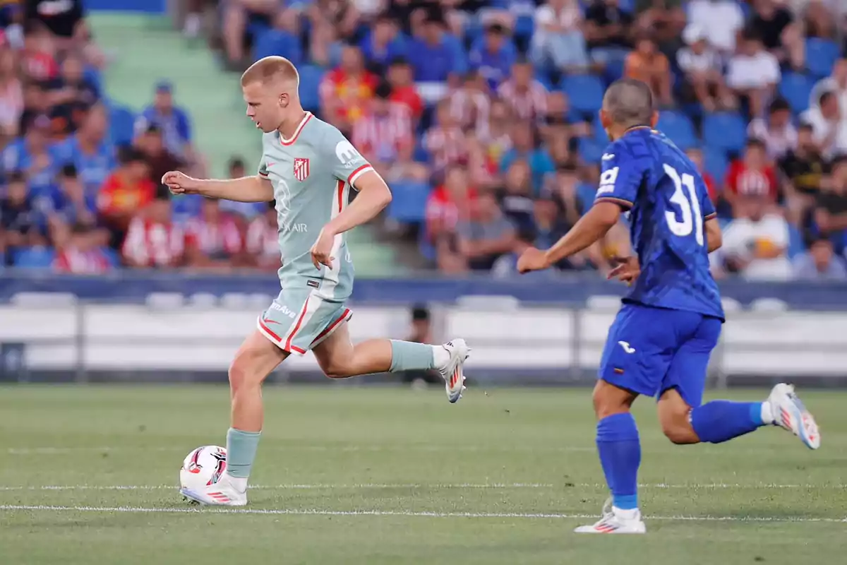 Two soccer players in action during a match, one in a blue uniform and the other in a gray uniform, in a stadium with spectators in the background.