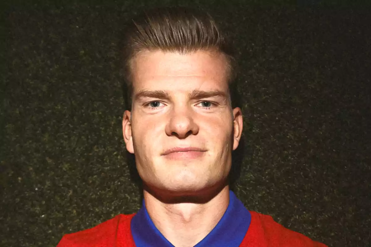 Portrait of a man with short blonde hair, wearing a red t-shirt with a blue collar, on a dark background.